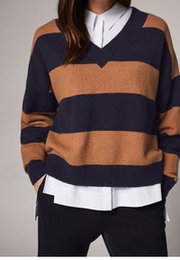 Pull en V marine et brun à rayures horizontales, fabriqué en tissu doux avec des poignets et un ourlet côtelés, superposé sur une chemise blanche à col.