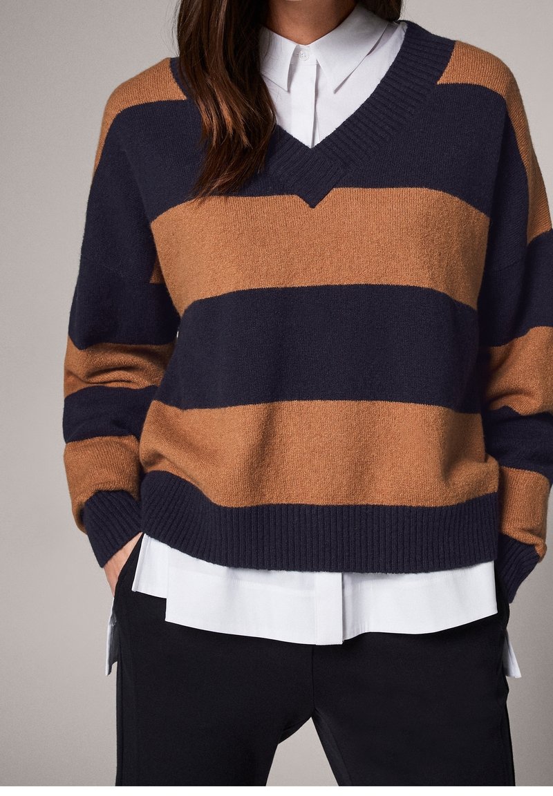 Pull en V marine et brun à rayures horizontales, fabriqué en tissu doux avec des poignets et un ourlet côtelés, superposé sur une chemise blanche à col.