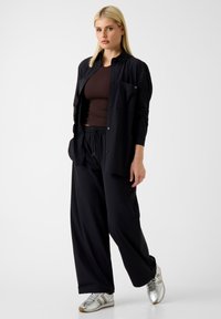 Zwart overhemd met knopen over een bruin shirt, gecombineerd met losse zwarte broek en zilveren sneakers. De stof lijkt glad en lichtgewicht.