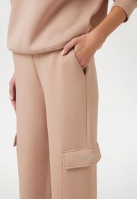 Šviesiai rožiniai sportiniai džinsai su lygia tekstūra. Rodo šoninė kišenė su uždengimu, papuošta prekės ženklo „GUESS“ kristalais.