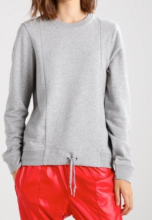 Vrouw die een lichtgrijze longsleeve trui met trekkoord draagt, gecombineerd met glanzende rode broek tegen een effen lichte achtergrond.