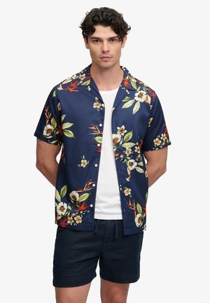 Uomo che indossa una camicia blu navy a maniche corte con bottoni e motivi floreali sopra una t-shirt bianca e pantaloncini blu navy, in piedi con le mani dietro la schiena su sfondo bianco.