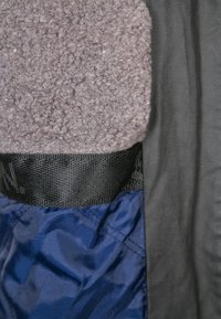 Gråt fleece materiale øverst, sort stof til højre, med blåt stof synligt nedenfor, sikret af en sort rem.