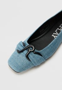 Džínová plochá obuv ve světle modré barvě s čtveratou špičkou, s detailním uzlem a černými kovovými akcenty. Hladká textura a minimalistický design.