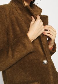 Hnědý kabát z vlněné směsi s vysokým límcem, zakončený stříbrným knoflíkem a tvořený měkkou, texturovanou látkou. Vykazuje přiléhavou siluetu.
