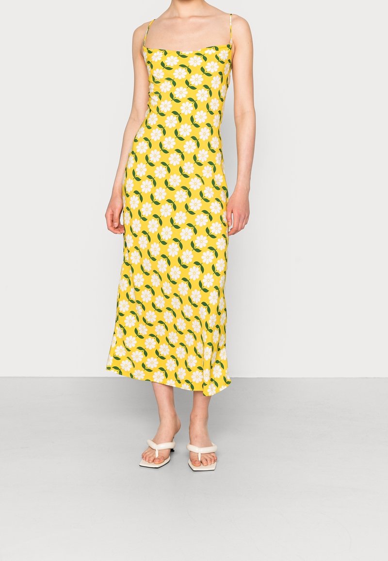 Femme portant une robe midi jaune avec un motif de fleurs blanches et de feuilles vertes et des sandales blanches à talons, debout devant un fond uni.