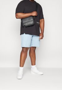 Mies, jolla on päällään musta Calvin Klein -paita, vaaleansiniset farkkuhousut, valkoiset tennarit ja joka kantaa mustaa olkitarraa.
