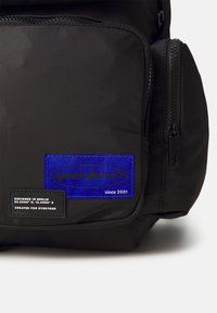 Black backpack made of durable fabric. Features a blue and black logo patch, zippered compartments, and textured finish.