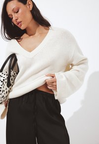 Una donna in un maglione bianco a maglia con scollo a V e pantaloni neri con cordoncino tiene una borsa a pois bianca e nera contro uno sfondo bianco puro.