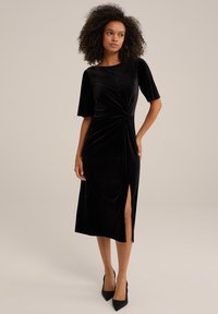 Black velvet midi dress with short sleeves, featuring a twisted front design and a thigh-high slit. Model stands in black high heels.