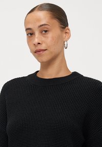Zwarte gebreide trui met een gestructureerd patroon, met een ronde hals en geribbelde manchetten. Gecombineerd met een model dat oorbellen met ringen draagt.
