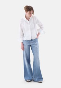 Weißes Langarm-Hemd mit Knopfleiste und abgerundetem Saum, kombiniert mit weit geschnittenen hellblauen Denim-Jeans. Das Outfit weist subtile, kontrastierende Texturen auf.