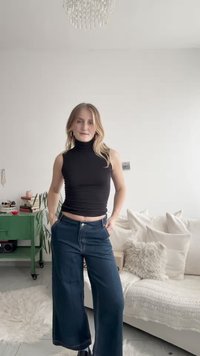 Svart ärmlös polotröja, högmidjade vida blå jeans, händerna i fickorna, texturen kontrasterar mot mjuka möbler i bakgrunden.