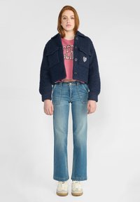 Veste cropped texturée bleu marine avec deux poches à l'avant et fermeture à boutons, associée à un sweat-shirt graphique rose et à un jean évasé bleu clair.
