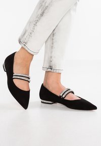 Bailarinas de ante negro con punta afilada. Presentan dos tiras adornadas con cristales y un detalle decorativo en el tacón. Llevadas con jeans de color claro.