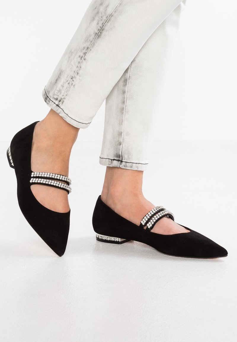 Bailarinas de ante negro con punta afilada. Presentan dos tiras adornadas con cristales y un detalle decorativo en el tacón. Llevadas con jeans de color claro.