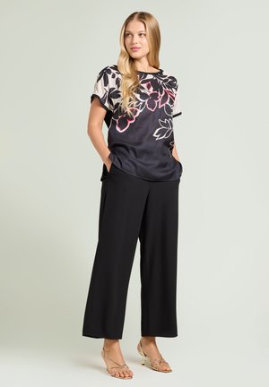 Kobieta stojąca z rękami w kieszeniach, ubrana w czarne spodnie o szerokich nogawkach, czarno-białą kwiecistą bluzkę z krótkim rękawem oraz złote sandały na paski.