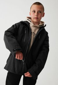Schwarze wasserdichte Jacke mit Reißverschluss und Kapuze, mit verstellbaren Bündchen und mehreren Taschen. Trägt man über einem beigefarbenen Hoodie.