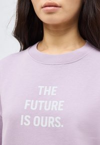 Νεαρός που φοράει ένα ανοιχτό μωβ φούτερ με λευκό κείμενο «ΤΟ ΜΕΛΛΟΝ ΕΙΝΑΙ ΔΙΚΟ ΜΑΣ» στο μπροστινό μέρος, φωτογραφία κομμένη στο ύψος του λαιμού.