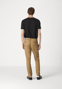Chinos de algodón beige con un corte ajustado y bolsillos traseros, combinados con una camiseta negra de manga corta, vistos desde atrás. Zapatillas negras con acentos blancos.