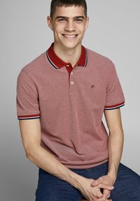 Rood en wit gestreept poloshirt met een rode kraag en blauwe-witte accentstrepen. Twee knopen bij de kraag. G gedragen met een blauwe spijkerbroek.