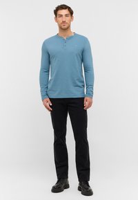 Turkusowa koszula henley z długim rękawem i trzema guzikami, zestawiona z czarnymi spodniami i czarnymi sznurowanymi butami. Prosty, teksturowany materiał.