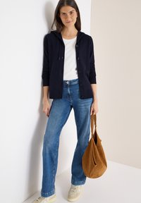 Felpa blu con bottoni, camicia bianca, jeans blu, sneakers beige, in mano una borsa in camoscio marrone, in piedi contro uno sfondo chiaro.