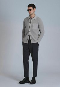 Light gray knit cardigan with a chest pocket, button closure, worn over a dark top and tapered navy trousers, complemented by black shoes and sunglasses.