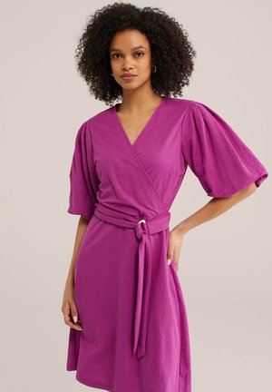 Lokkis juustega naine, kes kannab fuksia värvi ümber mähitavat kleiti, millel on õlgadest küünarnukkideni ulatuvad pundunud varrukad ja vöökohal seotud vöö.