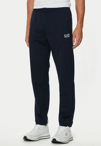 Uomo che indossa pantaloni tuta neri EA7 e sneaker bianche, in piedi contro uno sfondo chiaro e uniforme.