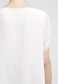 Camicia bianca con vestibilità ampia, caratterizzata da maniche corte trasparenti, tessuto morbido e un ampio scollo rotondo. Design minimalista senza pattern visibili.