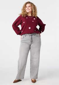 Gekrulde vrouw met een glimlach en handen op de heupen, gekleed in een bordeauxrood gebloemd sweater, lichtgrijze wijde jeans en luipaardprint flats.