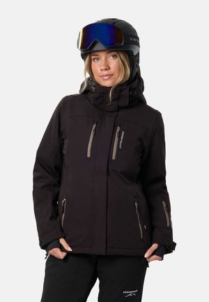 Une femme présente une veste et un pantalon de ski noirs, avec un casque noir et des lunettes de ski bleues, sur un fond uni.