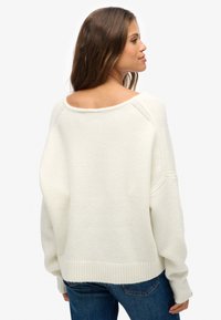 Witte oversized trui met een brede halslijn, geribbelde manchetten en zoom, en zachte, gestructureerde stof. Gecombineerd met een blauwe spijkerbroek.