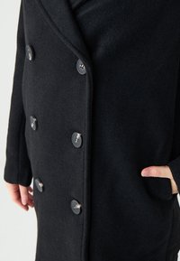 Schwarzer Wollmischmantel mit einem doppelreihigen Design, ausgestattet mit großen schwarzen Knöpfen und einer Seitentasche. Weicher, strukturiertes Material.