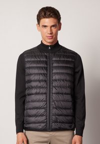 Svart puffer-väst med hög krage och framdragkedja. Har en kviltad textur och figursydd design, lagd över en svart tröja.
