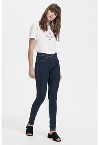 Jeans skinny azul oscuro de denim, combinados con una camiseta blanca con texto negro. Unos zapatos negros de tacón abierto completan el conjunto.