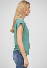 Turkis teksturert bluse med korte ermer, sammen med blå jeans. Sidemiljøvisning fremhever avslappet passform og subtile design detaljer.