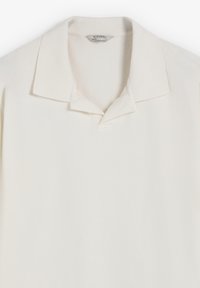 Polo de color blanco roto, con un cuello clásico y una textura suave. Detalle de costura sutil cerca del cuello. Etiqueta visible en el interior.