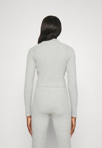 Persona con lunghi capelli scuri che indossa un maglione aderente grigio chiaro a maniche lunghe e pantaloni abbinati, in piedi con le spalle rivolte verso la telecamera su uno sfondo neutro.