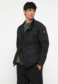 Black insulated jacket with a corduroy collar, two front pockets, and snap closures. Worn over a green top, featuring a branded patch.