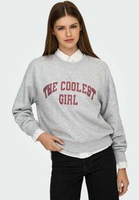 Hall hallipluus, millel on paksud punased tähed "THE COOLEST GIRL". Omadused on ümarsuu, pikad varrukad ja lahtine lõige valge kraega särgi kohal.