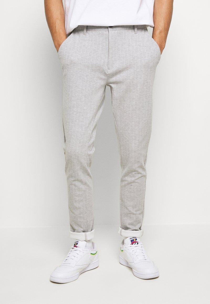 Homme portant un pantalon slim gris clair à fines rayures avec des revers et des baskets blanches Reebok, debout les mains dans les poches.