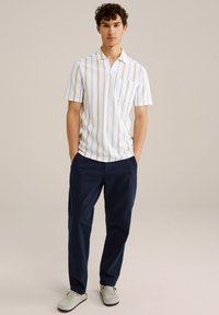 Camisa de manga corta con rayas verticales azules y beige, base blanca, y un bolsillo en el pecho, combinada con pantalones azul marino y zapatos gris claro.
