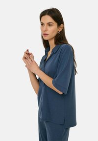 Mujer con cabello largo y oscuro, que lleva una camisa azul de manga corta y cuello suelta y pantalones a juego, con las manos suavemente entrelazadas cerca del rostro, con fondo neutro.