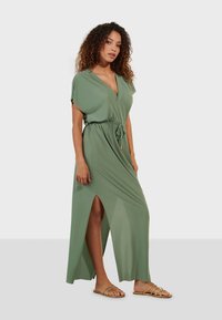 Robe maxi verte avec des manches courtes, décolleté en V, taille à cordon, fentes sur les côtés et texture lisse ; associée à des sandales dorées à lanières.
