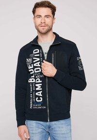 Felpa con zip navy e testo grafico bianco e blu, dotata di taschino sul petto e polsini a costine. Tessuto morbido e vestibilità casual.