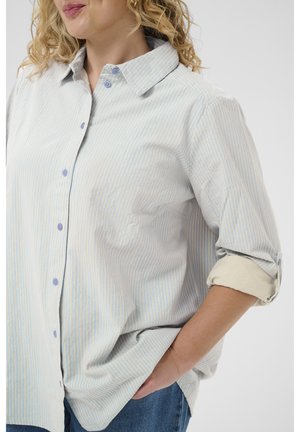 Frau mit lockigem blondem Haar trägt ein hellblau-weiß gestreiftes Hemd mit hochgekrempelten Ärmeln und blaue Jeans, Hand in der Tasche.