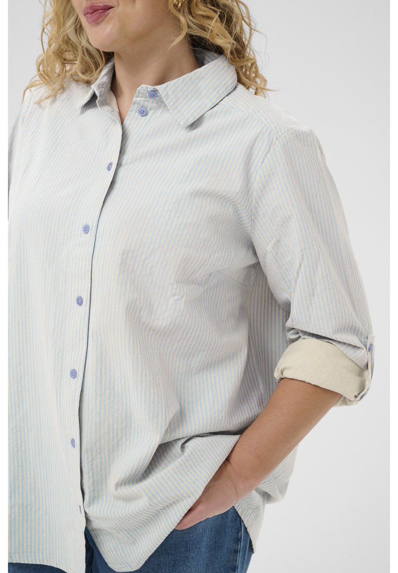 Femme aux cheveux blonds bouclés porte une chemise à rayures bleu clair et blanc, boutonnée, aux manches retroussées, et un jean bleu, main dans la poche.