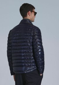 Doudoune marine avec une texture lisse et brillante, présentant un matelassage horizontal et un col. Porte par une personne, vue de dos.